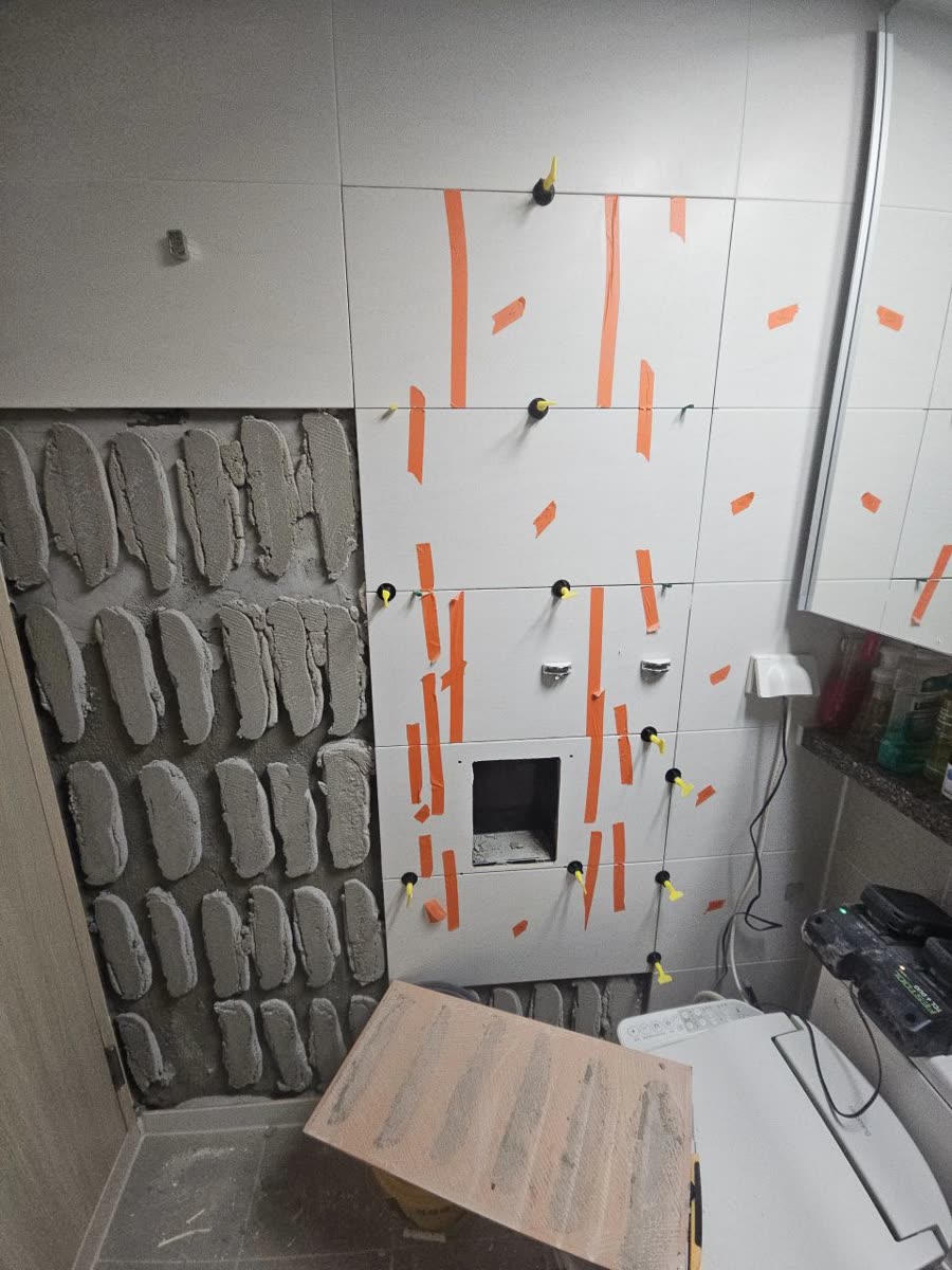 욕실 벽 한쪽 타일을 떼어낸 자리에 접착 자국이 남아 있고 옆에 새 타일이 임시로 붙은 시공 중 모습